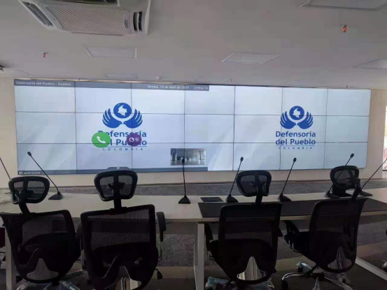 Conference room with several ergonomic chairs around a long table; a wall of large video screens displays the Defensoría del Pueblo Colombia logo and microphones on the desk.