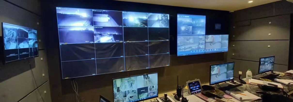 Security operations room with numerous wall-mounted monitors showing live CCTV feeds and a desk full of laptops and control gear in use.