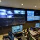 Security operations room with numerous wall-mounted monitors showing live CCTV feeds and a desk full of laptops and control gear in use.