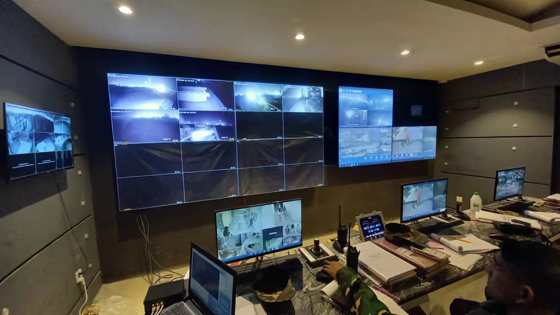 Security operations room with numerous wall-mounted monitors showing live CCTV feeds and a desk full of laptops and control gear in use.